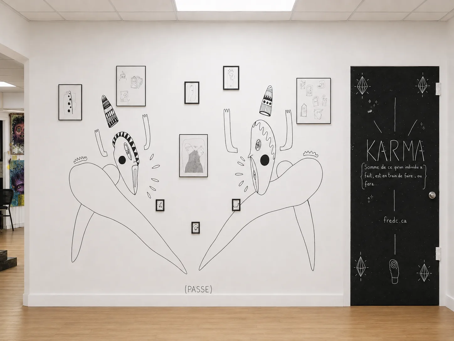 Mur de l'installation KARMA avec cinq dessins encadrés entourés de créatures tracées en noir, et une porte noire portant le mot KARMA et sa définition