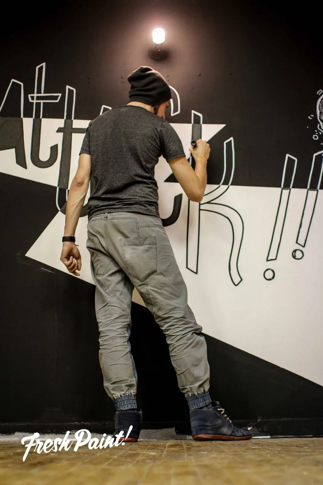 FREDC, de dos et coiffé d'un bonnet, trace au feutre le logo mural « ART ATTACK !! » sur le mur noir et blanc de la galerie